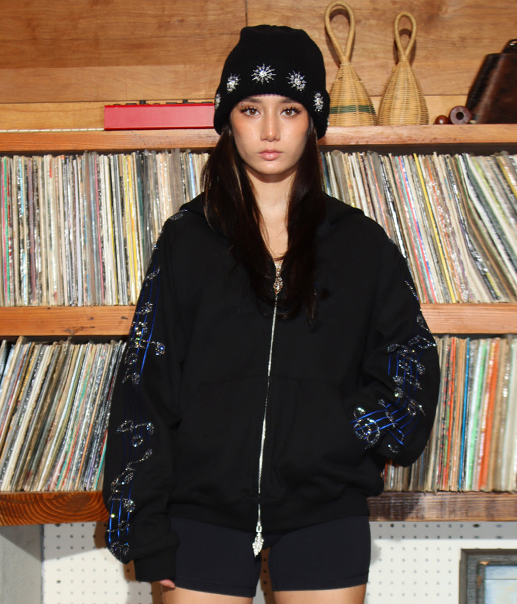 Person wearing a black jacket with blue heart design and matching beanie in front of a record collection.